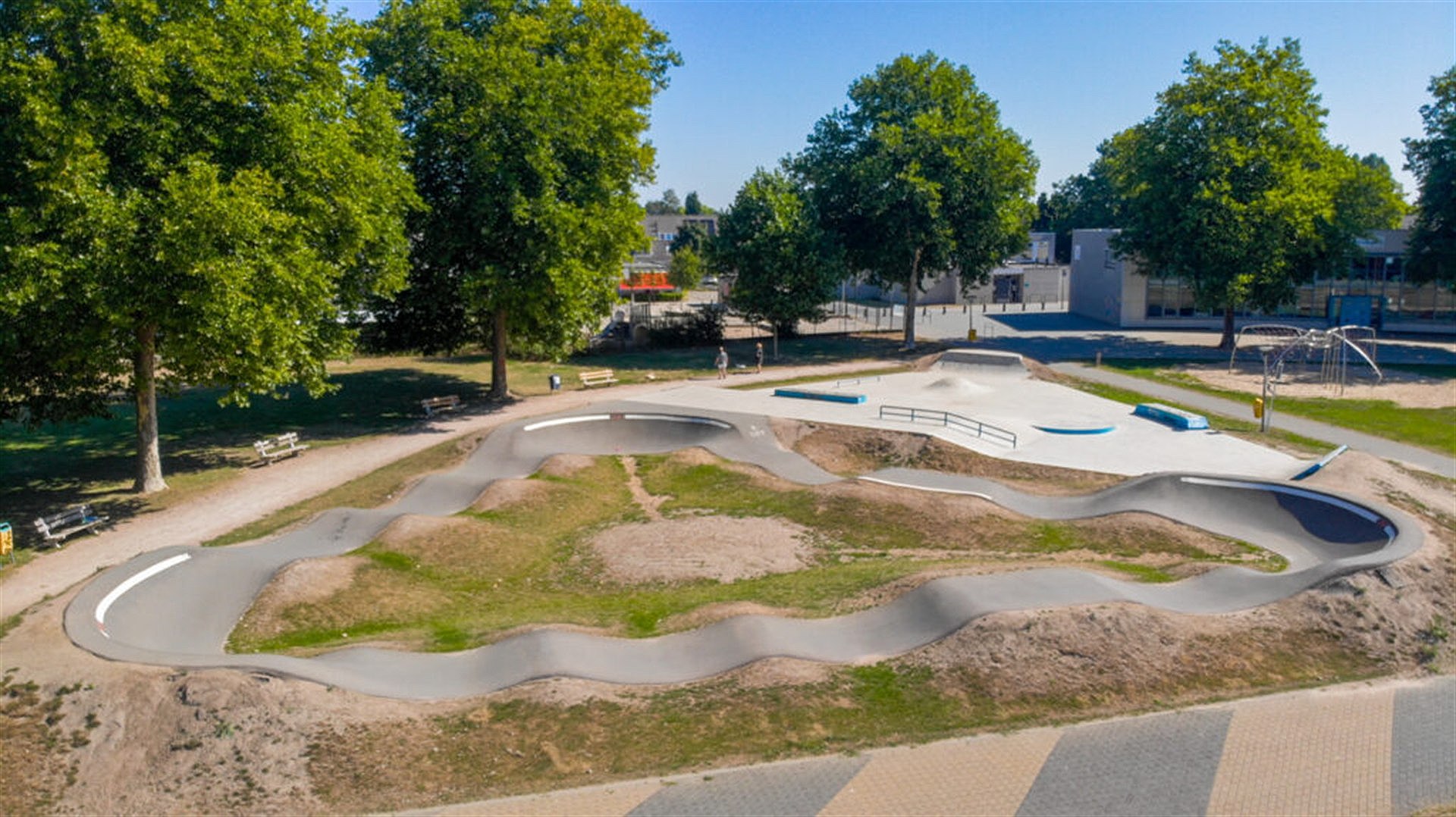 Asphalt pumptrack with looping rollers and berms, beside a concrete skate area in a public park