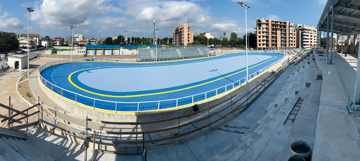 Banked speed track oval with blue surface and yellow lane markings, grandstand terracing in the foreground